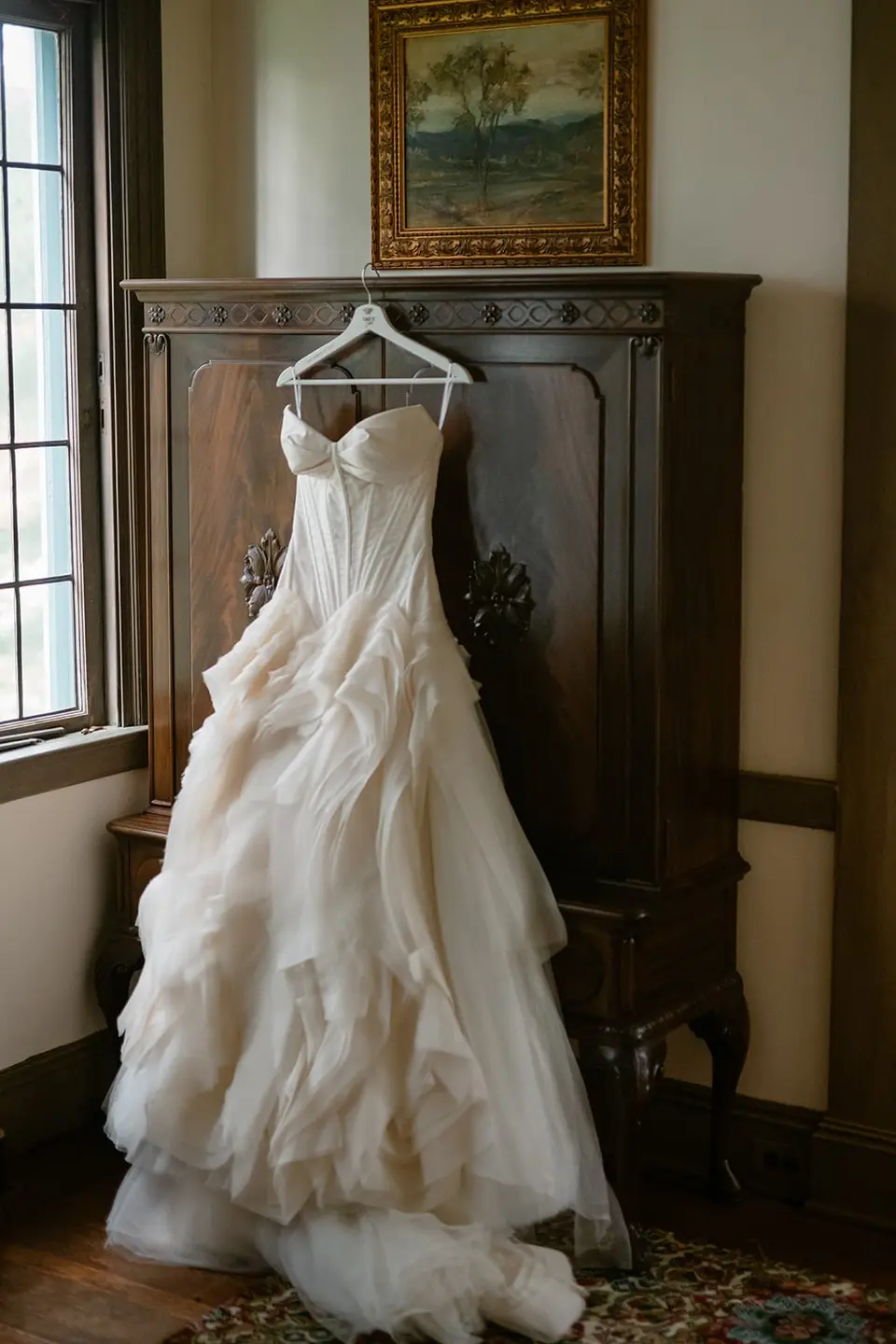 The ruffled wedding gown hanging from a carved wooden wardrobe beneath an oil painting.