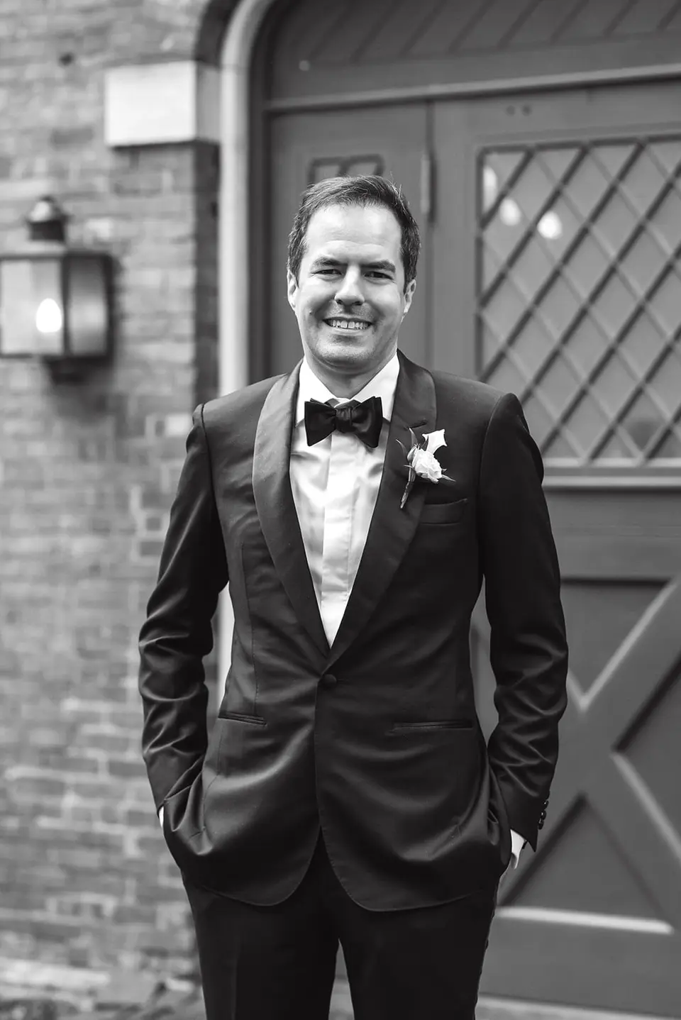 The groom in a tuxedo stands smiling before an arched wooden door. Black and white.