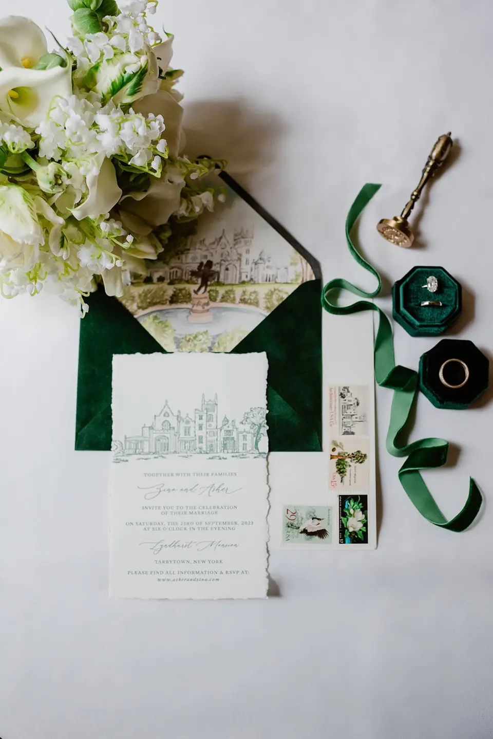 The invitation suite with a drawing of the mansion, a green velvet ring box, a wax seal and green ribbon beneath white flowers.