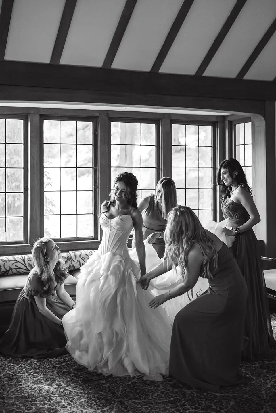 In a room with leaded windows, the bridesmaids gather round the bride to help with her gown. Black and white.
