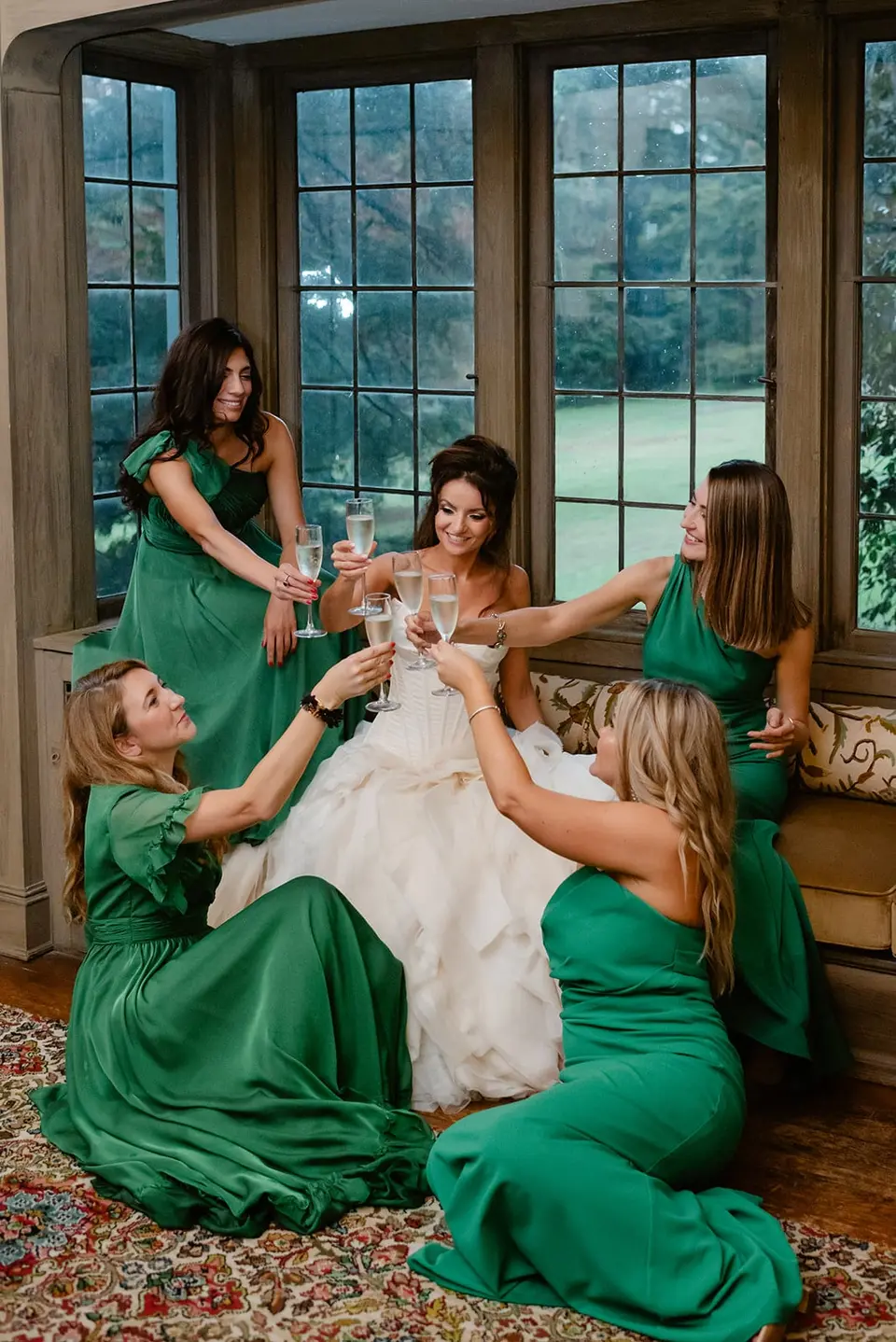 The bridesmaids in emerald raise their glasses to the bride on a window seat.