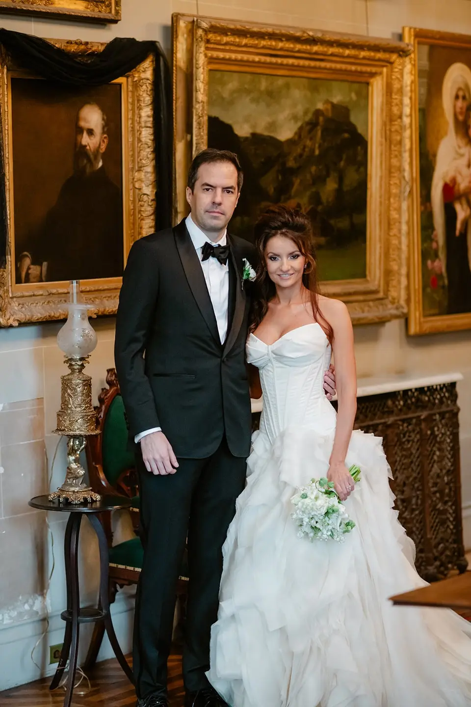 The bride and groom in a room hung with gilt-framed oil paintings.