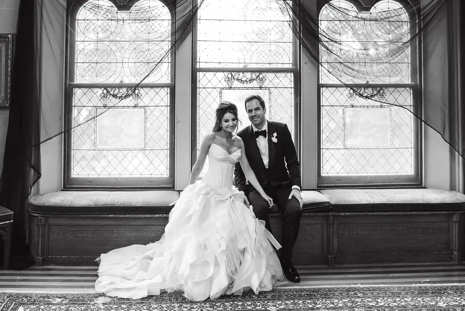 The bride and groom sit on a window seat beneath tall arched windows. Black and white.