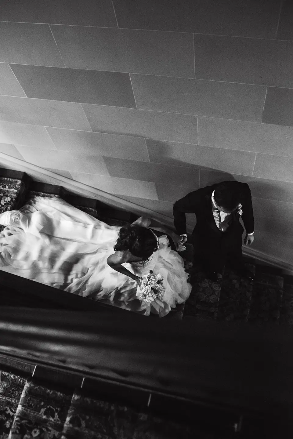From above, the bride and groom on a curving staircase, her train spread across the steps. Black and white.