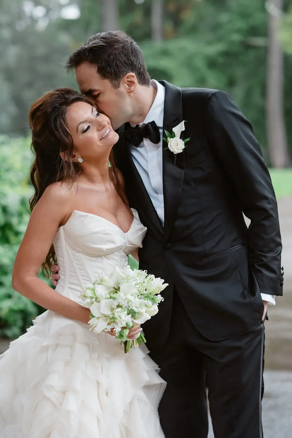 The groom kisses the bride's temple as she smiles, holding white flowers.
