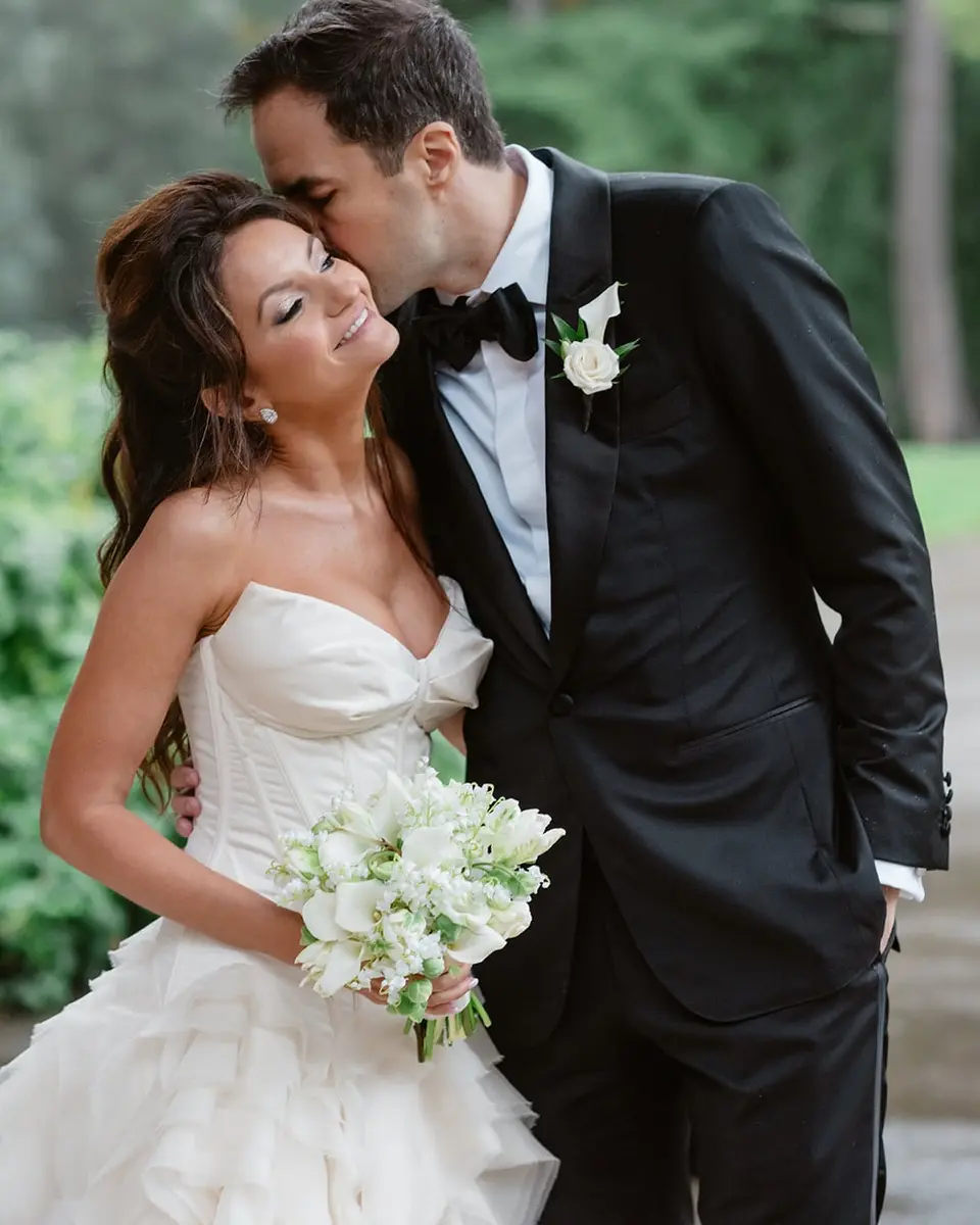At Lyndhurst Mansion, the groom kisses the bride's temple as she smiles, holding white flowers.