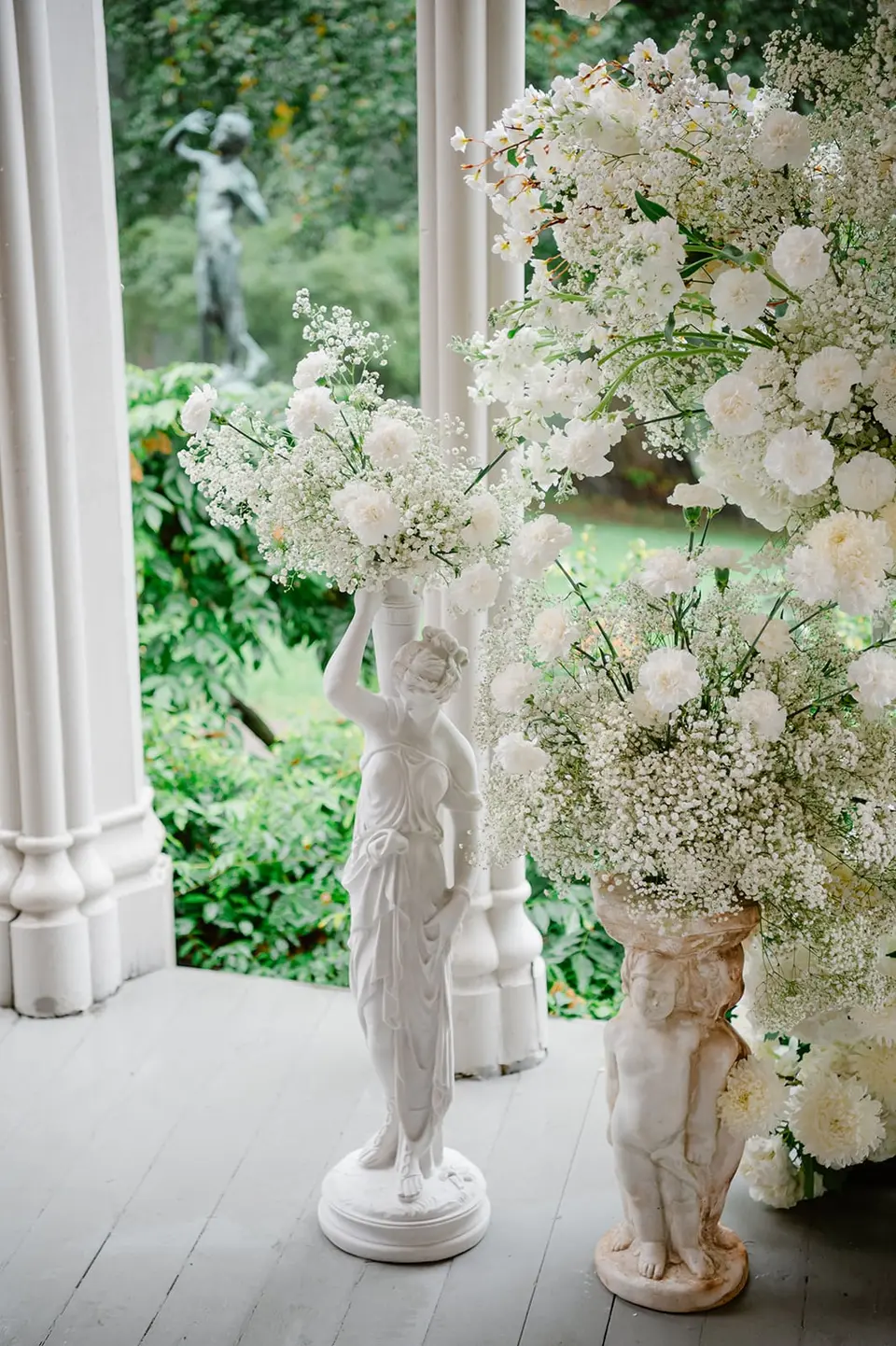 A white classical statue and an urn of white flowers on the veranda, the garden beyond.