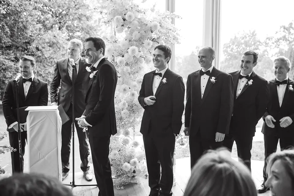 The groom laughs with the officiant and his groomsmen at the ceremony. Black and white.
