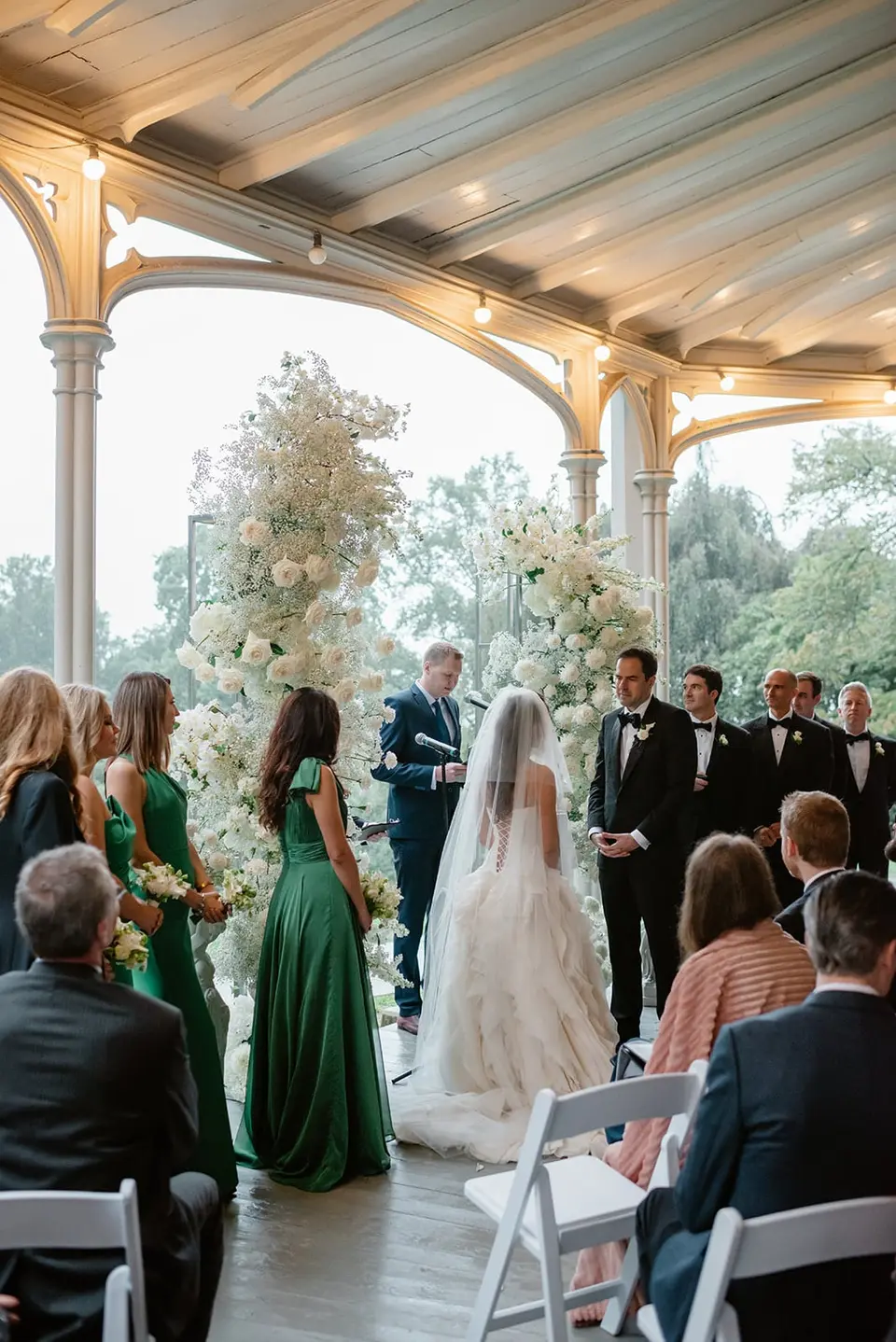 The ceremony on the veranda, bridesmaids in emerald and the couple beneath towers of white flowers.