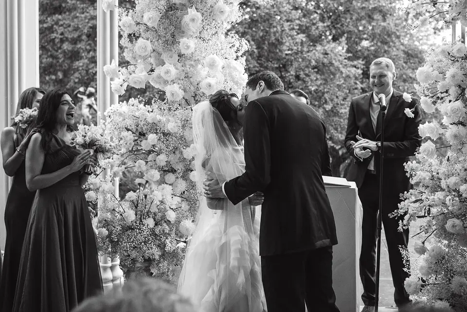 The bride and groom kiss between columns of white flowers as a bridesmaid laughs. Black and white.