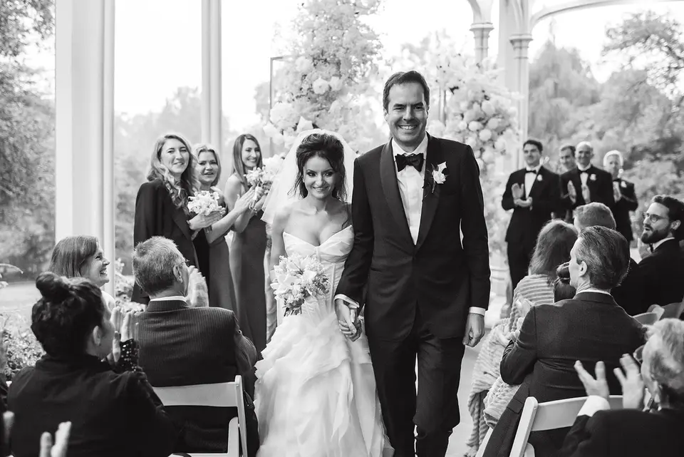 The bride and groom walk back up the aisle, married, as guests applaud. Black and white.
