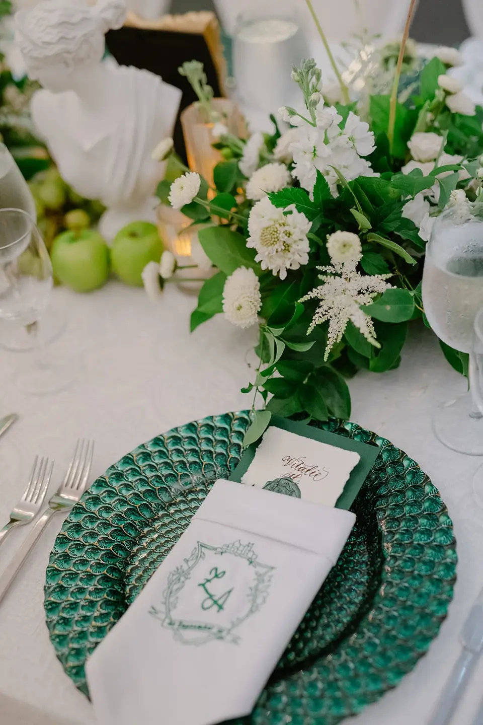 A place setting on an emerald glass charger, a monogrammed napkin and green apples among white flowers.
