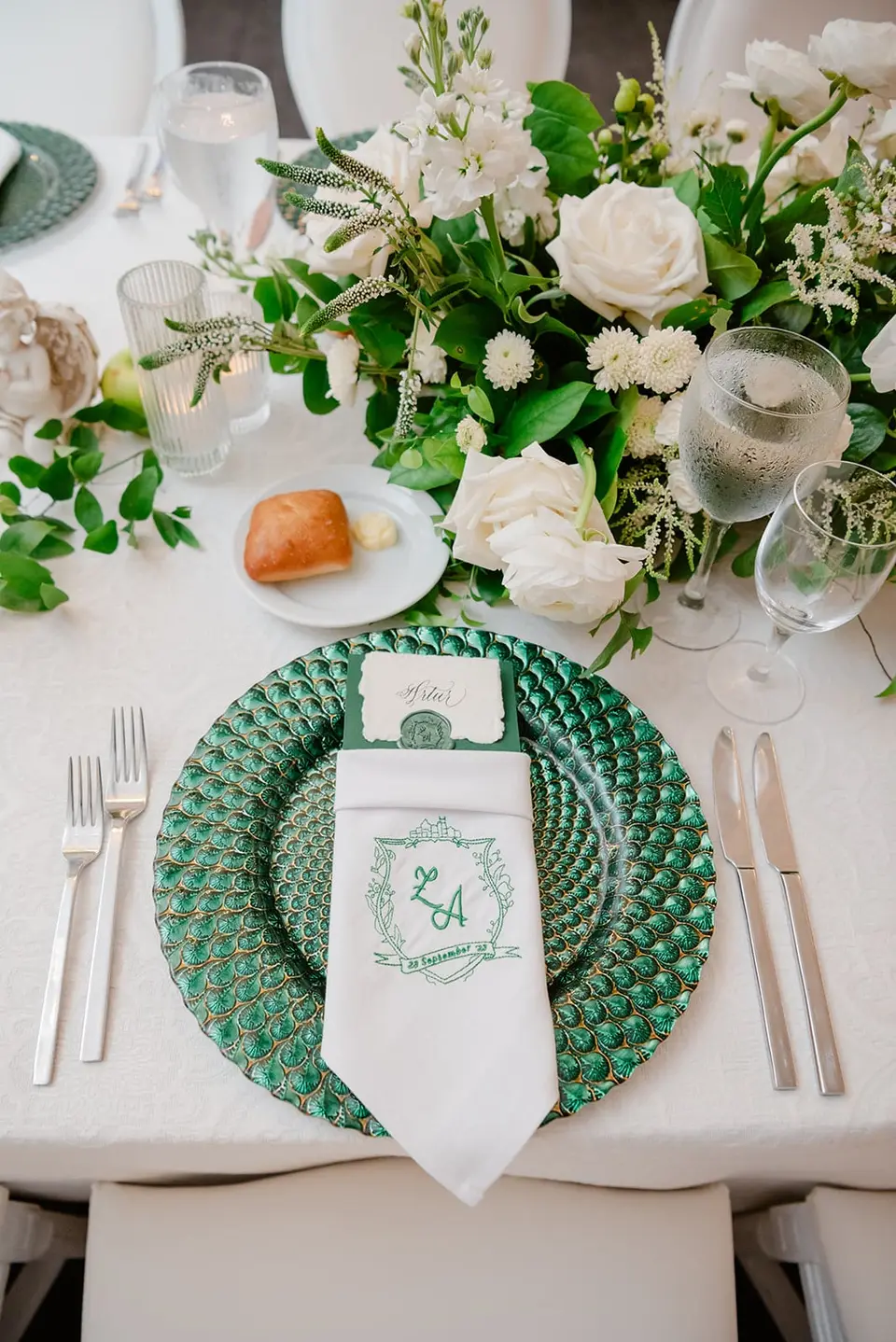 A place setting with a monogrammed napkin on an emerald charger, white roses and greenery across the table.