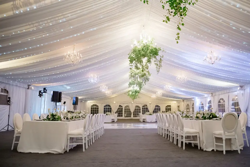 The tent set for dinner, long white tables beneath draped fabric, crystal chandeliers and hanging greenery.