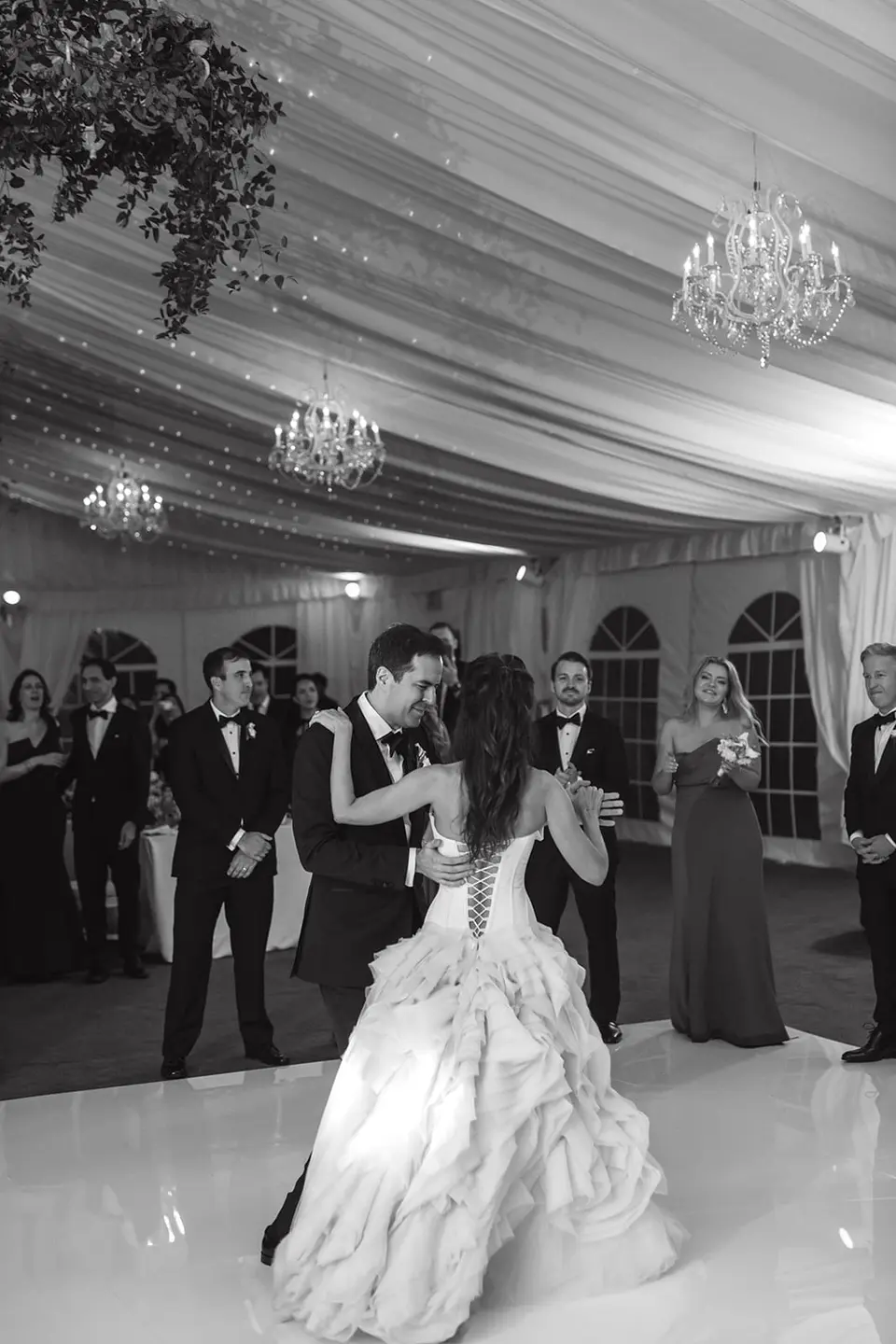The bride and groom take the floor for their first dance, guests watching. Black and white.