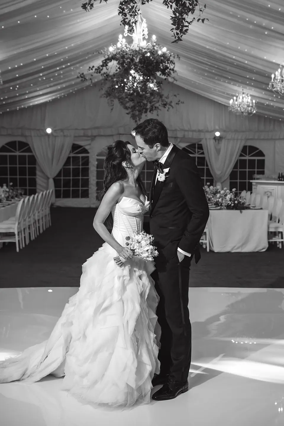 The couple kiss on the white dance floor beneath the chandeliers. Black and white.