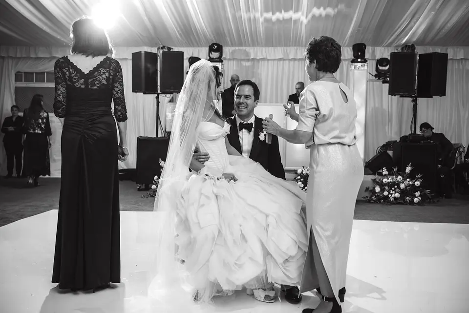 The bride and groom sit on chairs in the middle of the dance floor, family standing around them. Black and white.