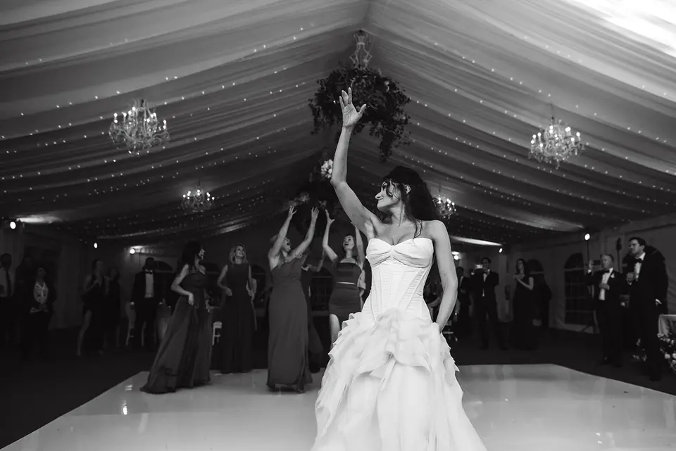 The bride raises her arm on the dance floor, the tent lights behind her. Black and white.