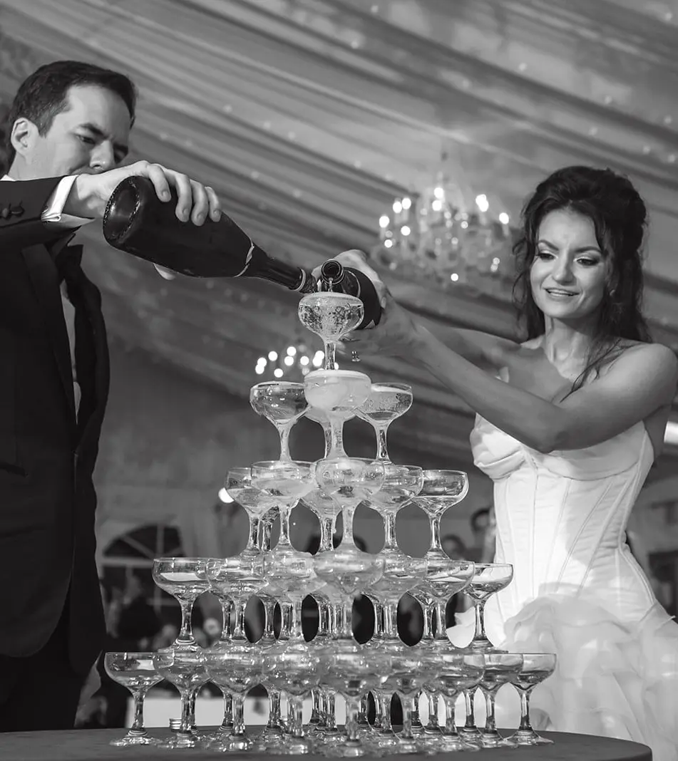 The groom pours champagne over a tower of coupes as the bride steadies a glass. Black and white.