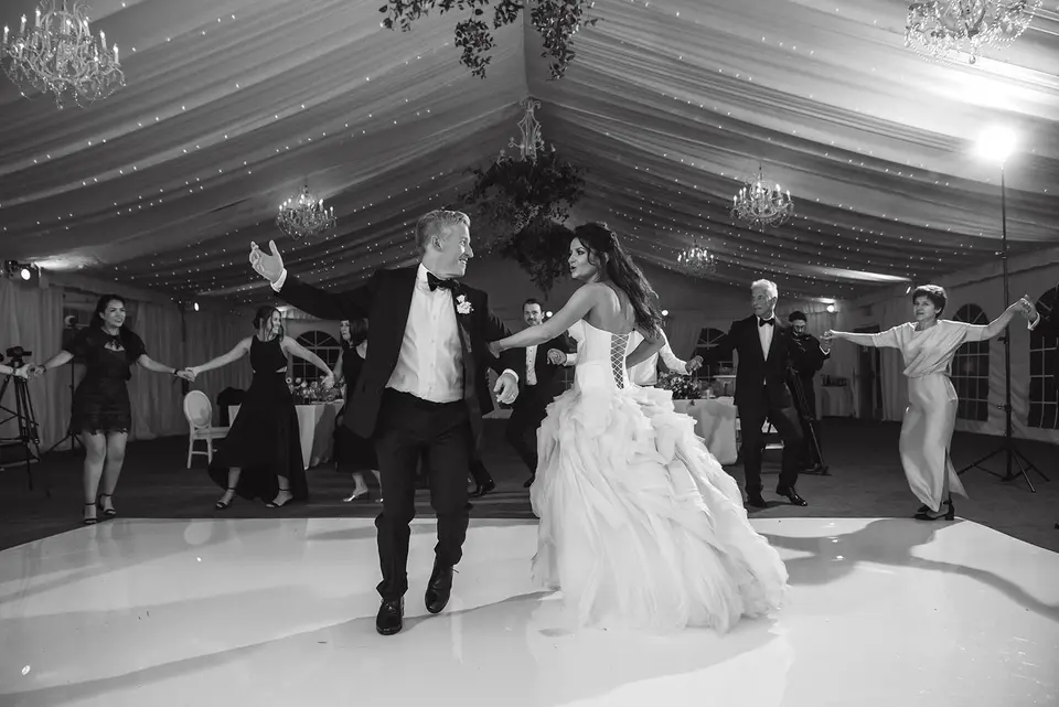 The bride and groom dance across the white floor, guests dancing behind them. Black and white.