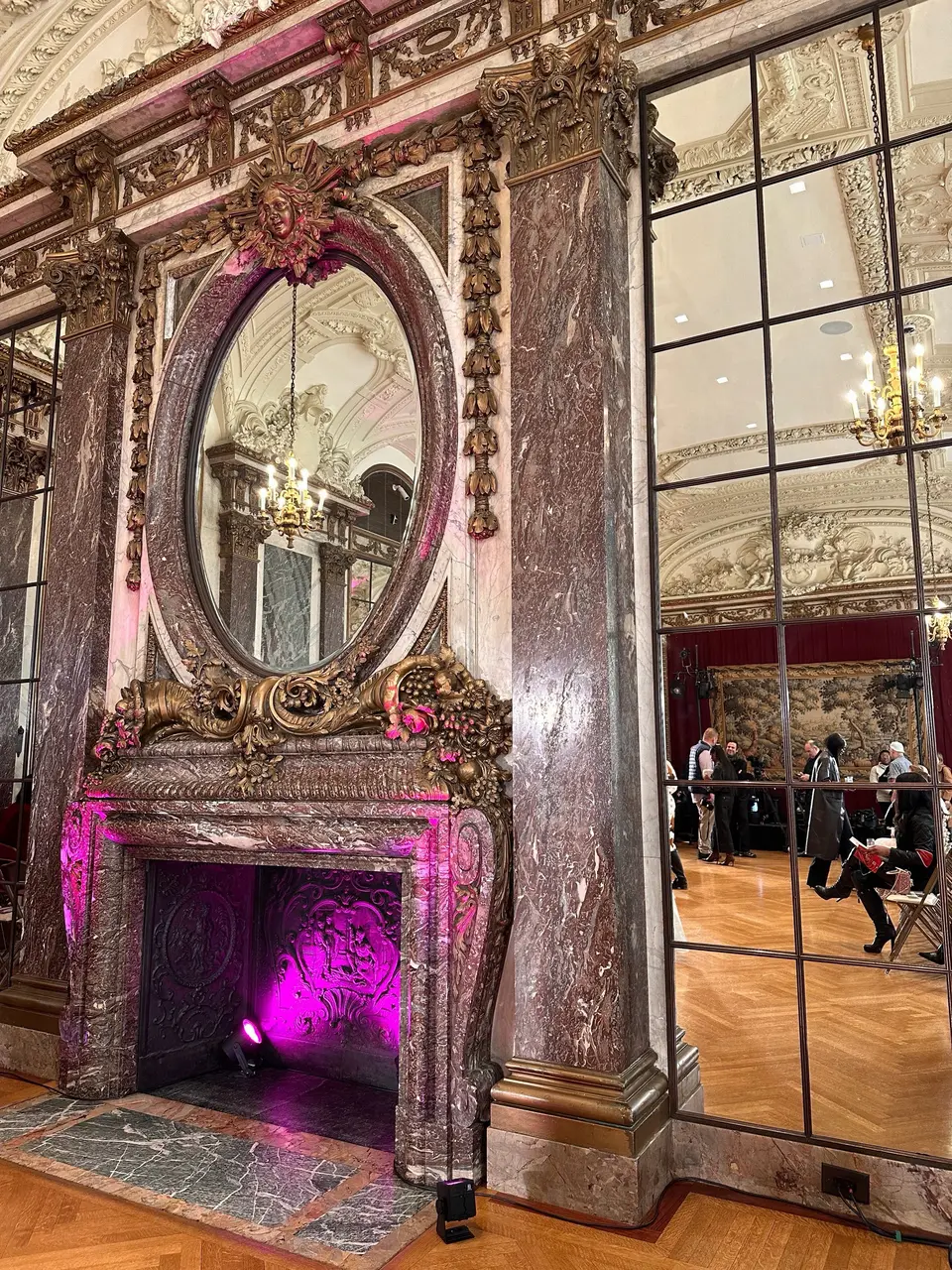 A gilded oval mirror above a fireplace of rouge marble, rich carved ornament around it and pink light glowing in the hearth.