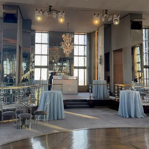 In the Rainbow Room, small chandeliers hang before a mirrored wall and tall windows, light blue cocktail tables set around a raised stage and the round parquet dance floor.
