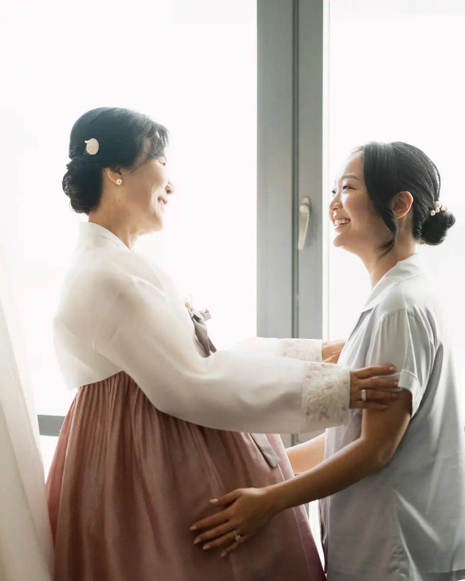 By a tall window, a woman in a white and rose hanbok holds the bride's arms and smiles at her, the gown hanging beside them.