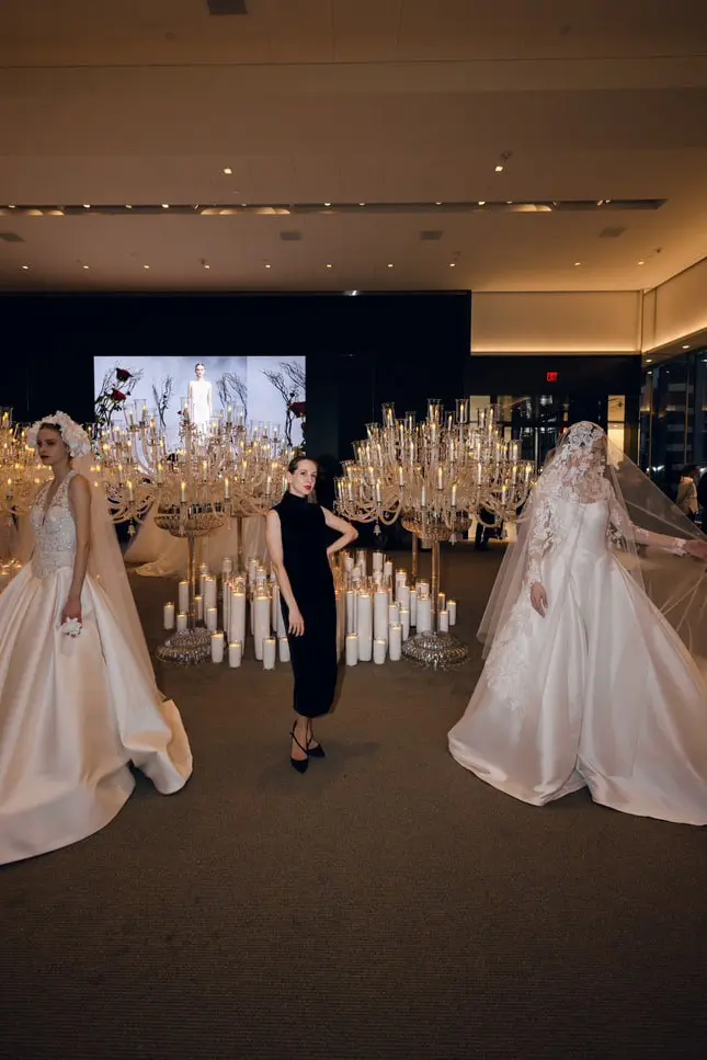 Olha Barabash, in a black dress, stands between tall candelabras of candles and two models in bridal gowns at New York Bridal Fashion Week.