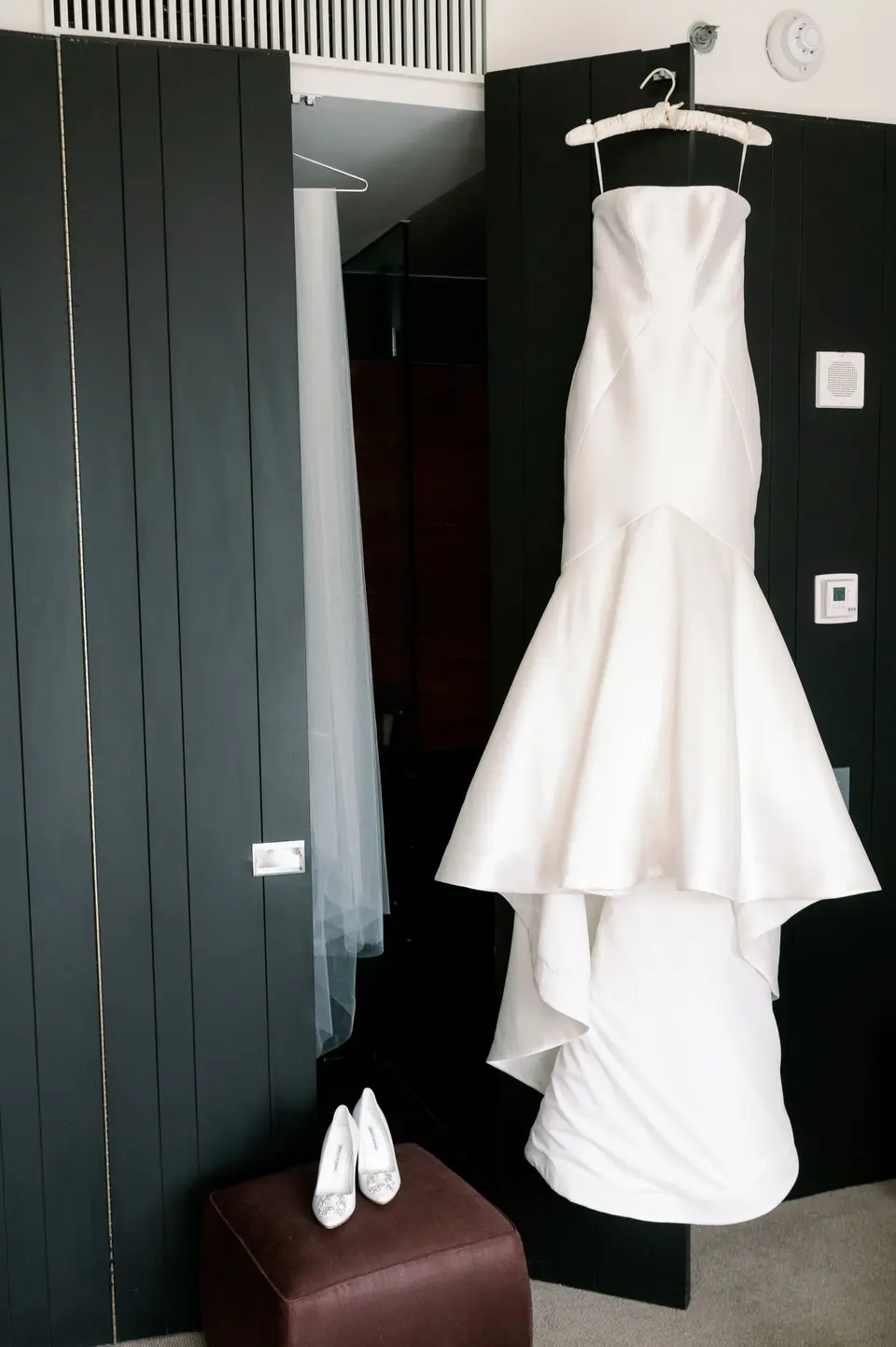The strapless wedding gown hanging in a dark-panelled hotel room, white shoes on a stool beneath it.