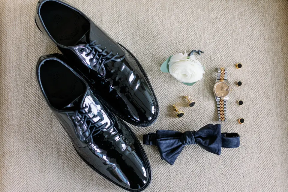 The groom's patent leather shoes laid out with a navy bow tie, a watch, cufflinks and a white boutonniere.
