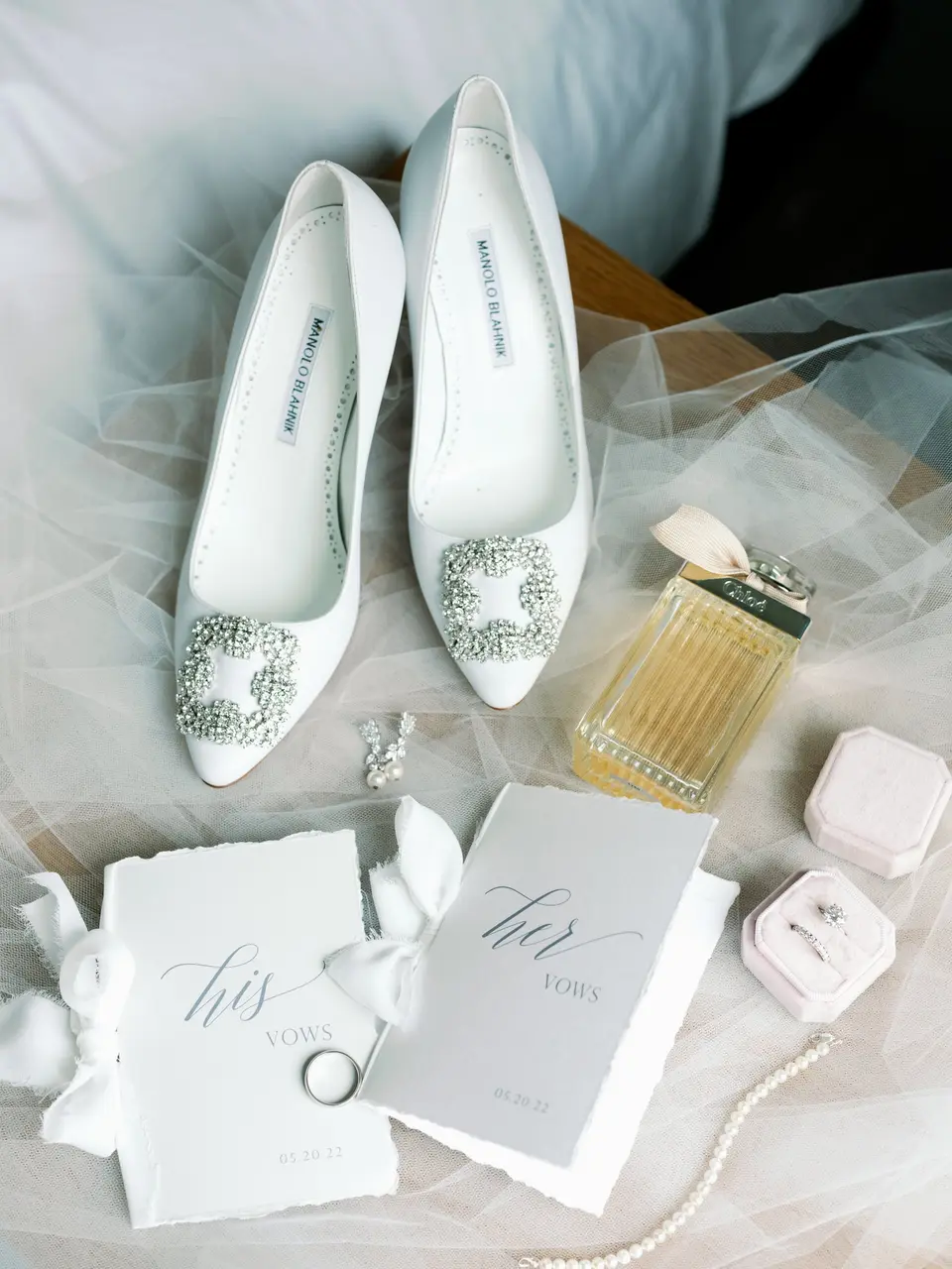 White satin heels with jeweled buckles, his and hers vow books, a ring box and perfume laid on tulle.