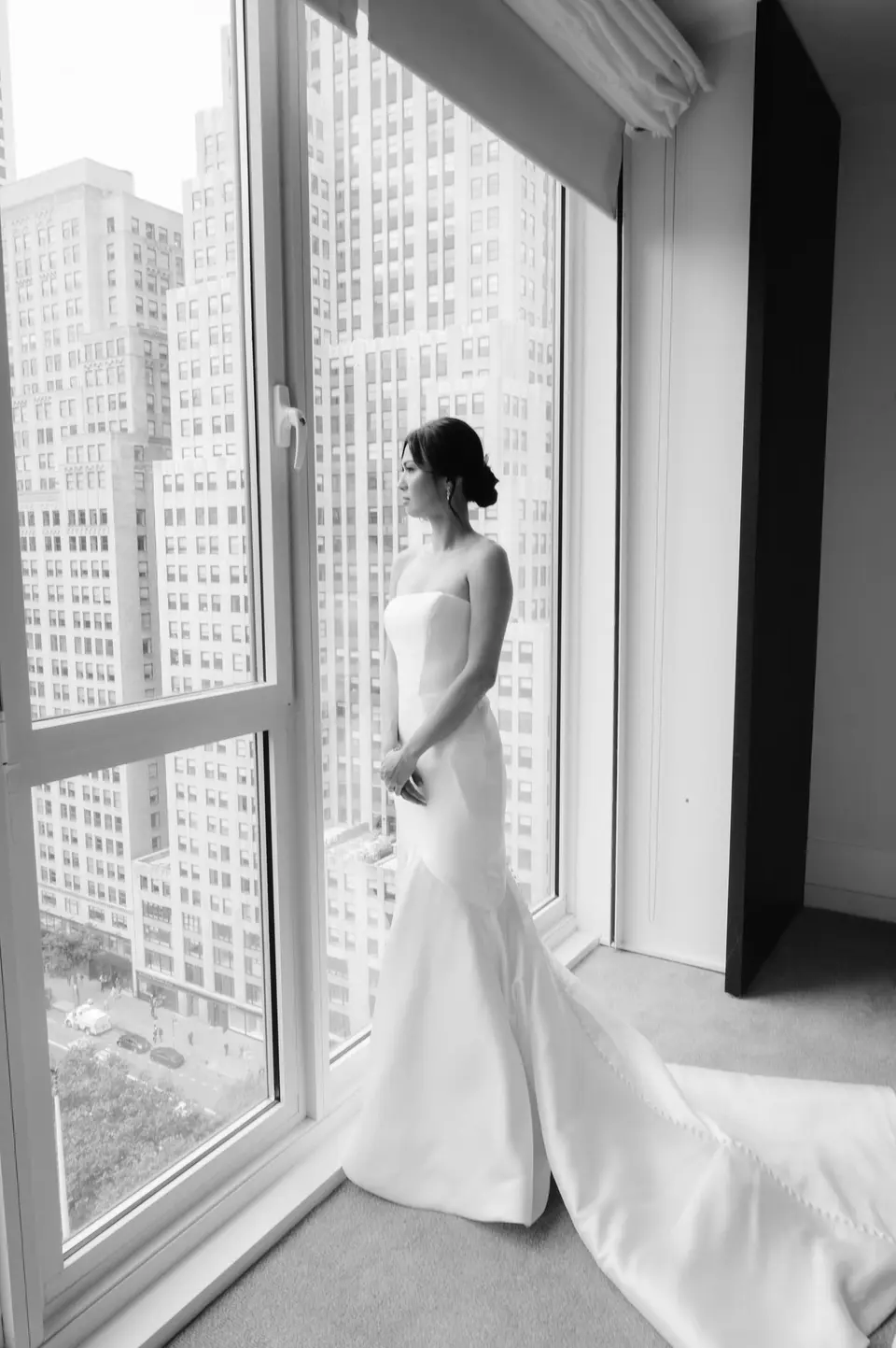 The bride stands at a tall window looking out over Midtown towers. Black and white.