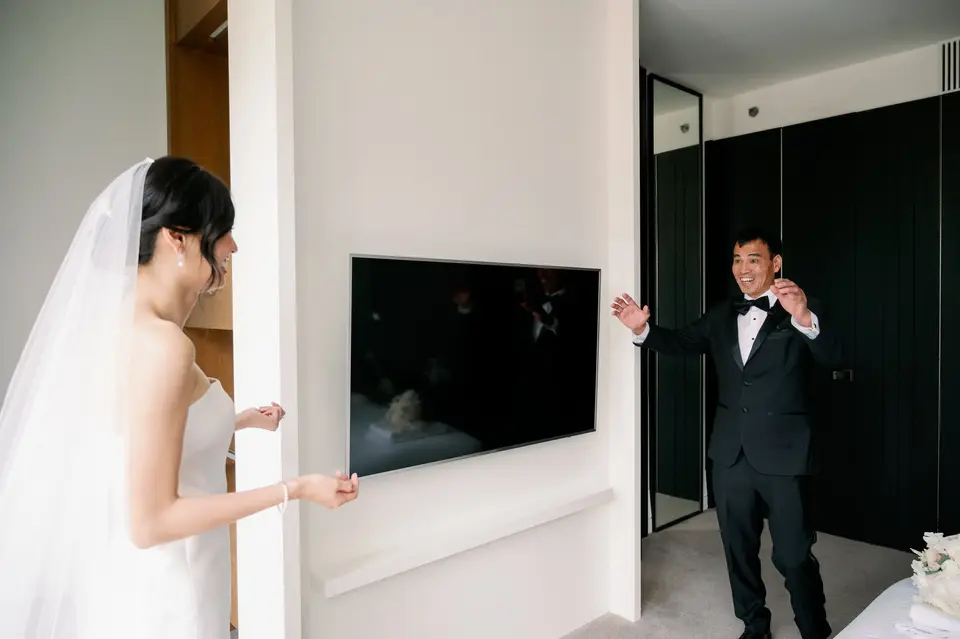 The bride peers around a wall at the groom, who throws up his hands in delight.