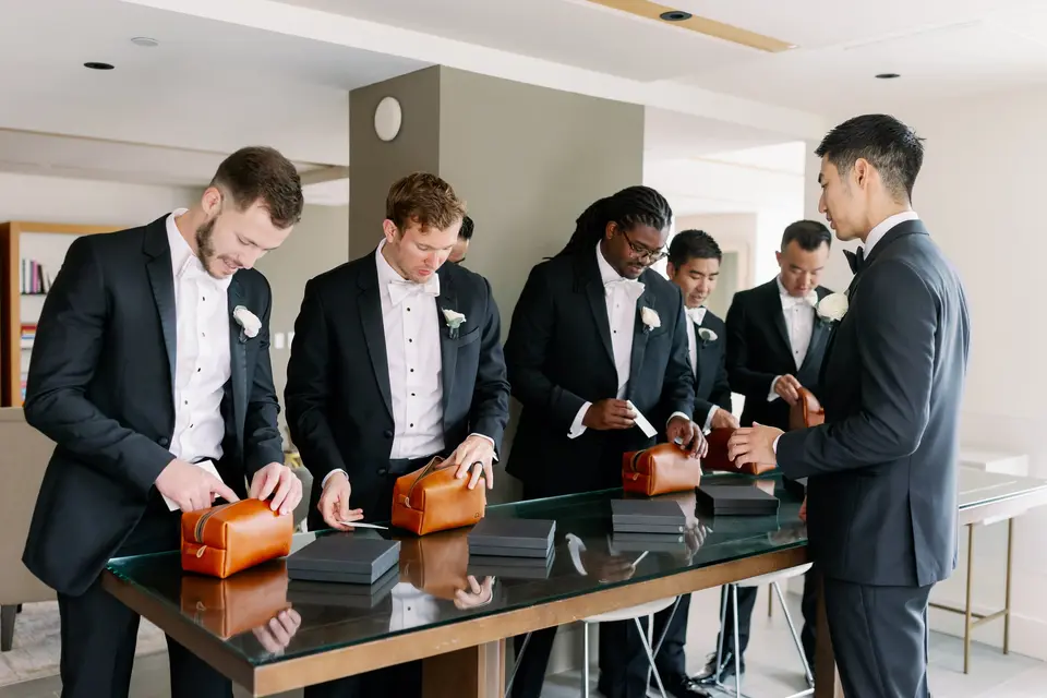 The groomsmen open gifts in leather cases at a glass table.