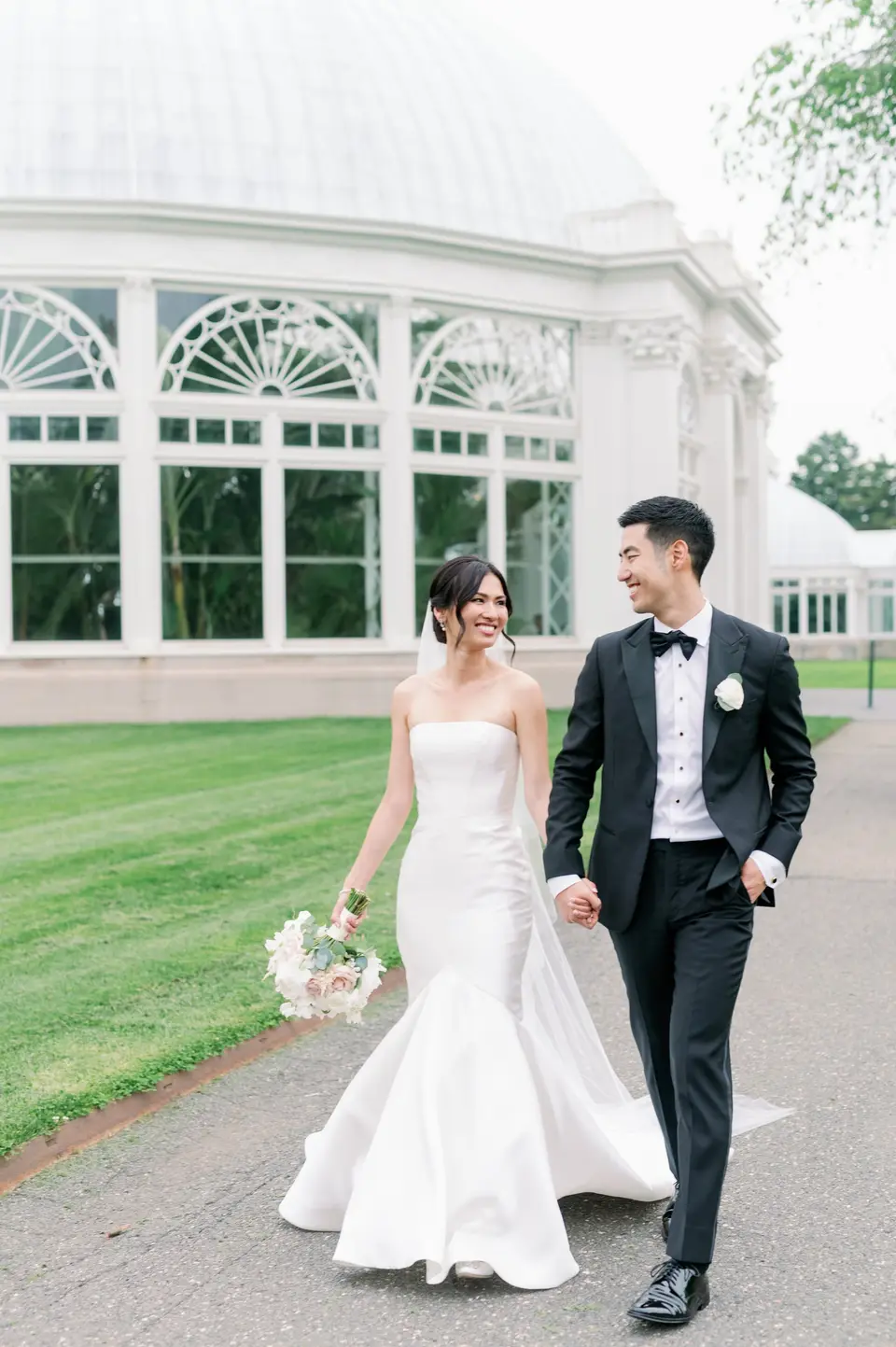 The bride and groom walk hand in hand in front of the glass conservatory at the Botanical Garden.