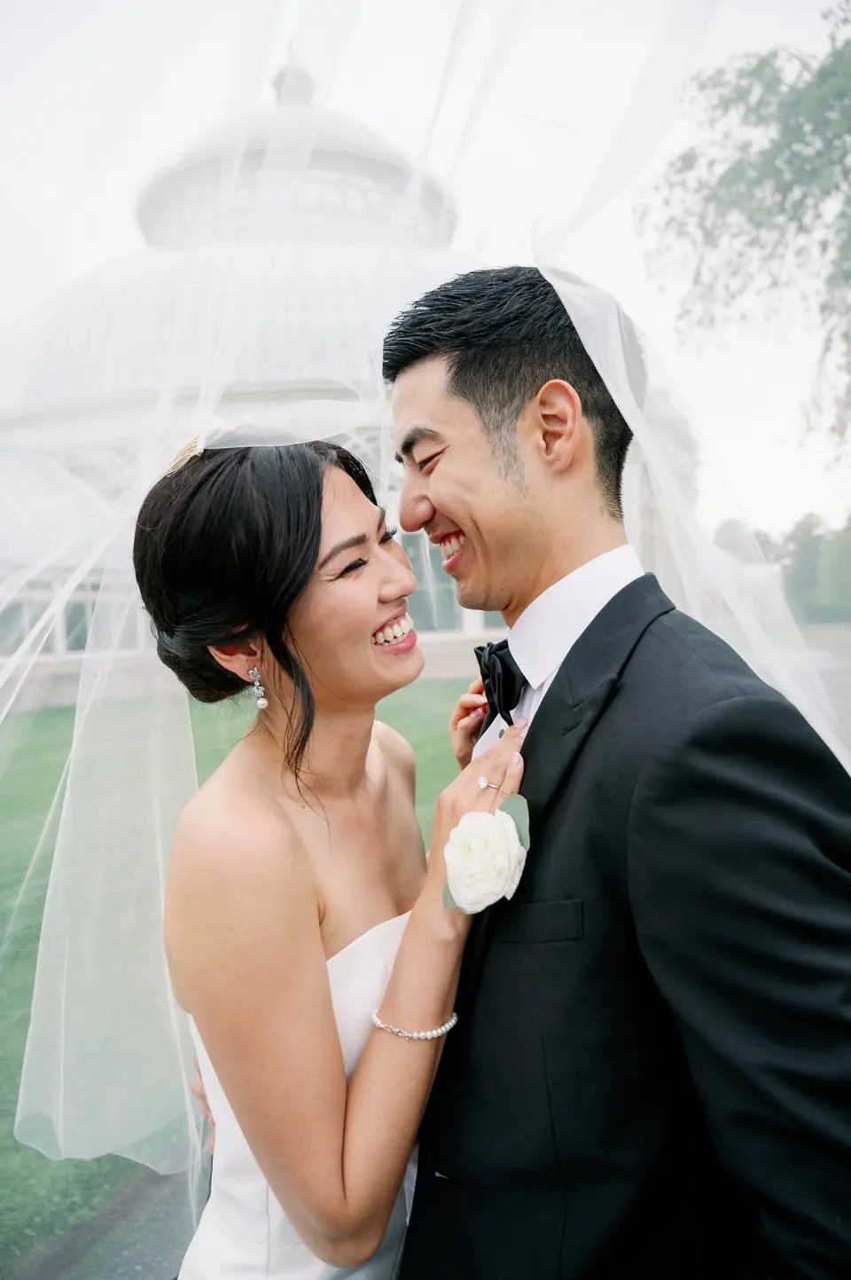Beneath her veil, the bride laughs with the groom, the conservatory dome behind them.