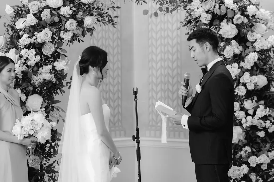 The groom reads his vows beneath an arch of white flowers. Black and white.
