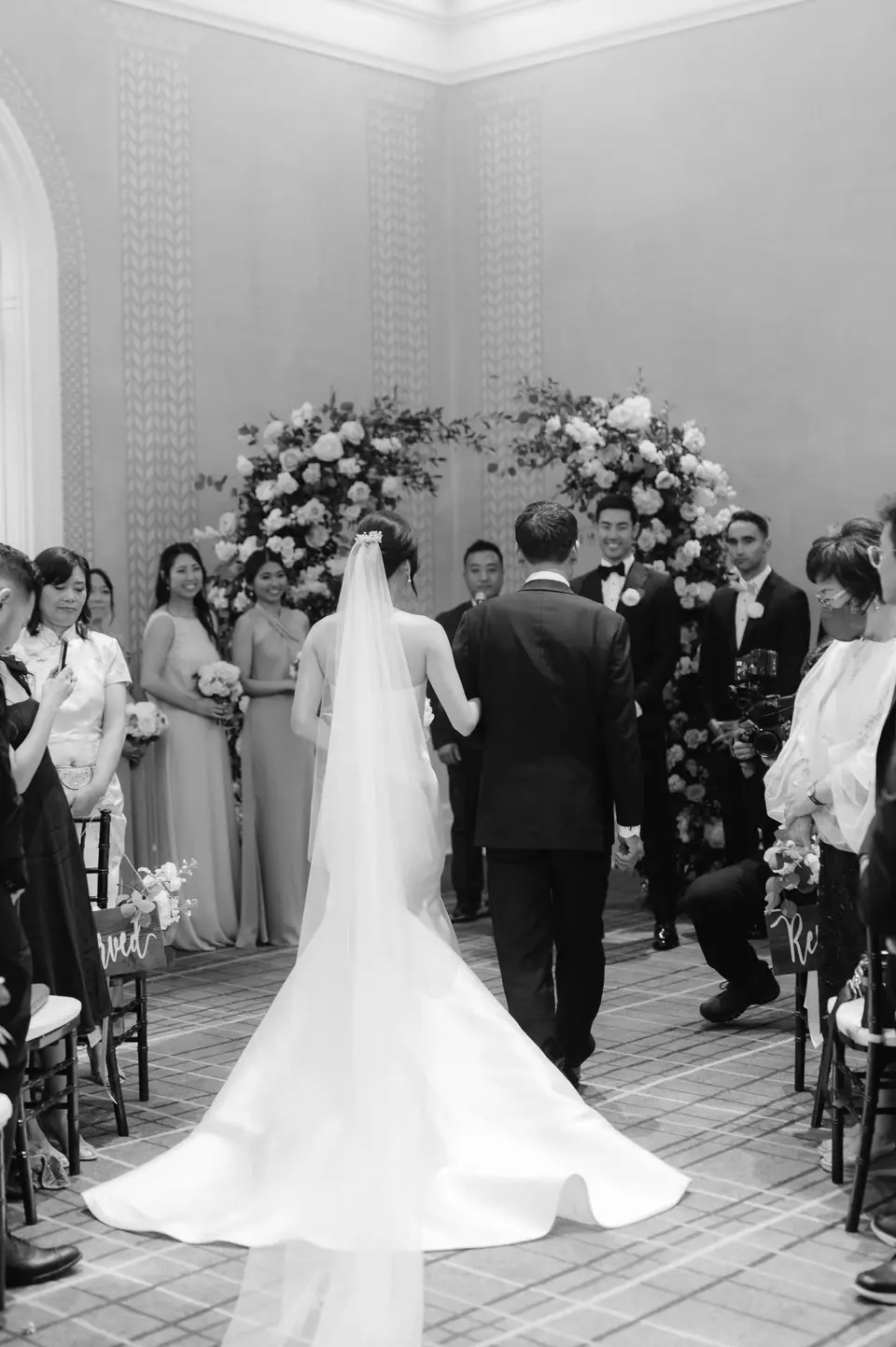 Seen from behind, the bride walks down the aisle on her father's arm. Black and white.
