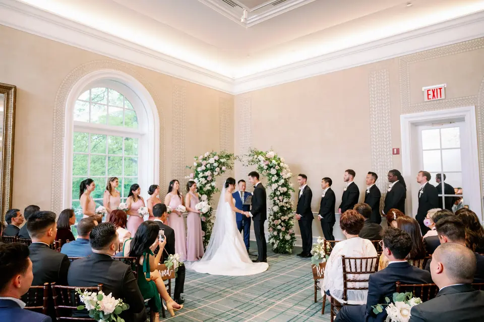 The ceremony in a room with tall arched windows, the wedding party standing on either side of the couple.
