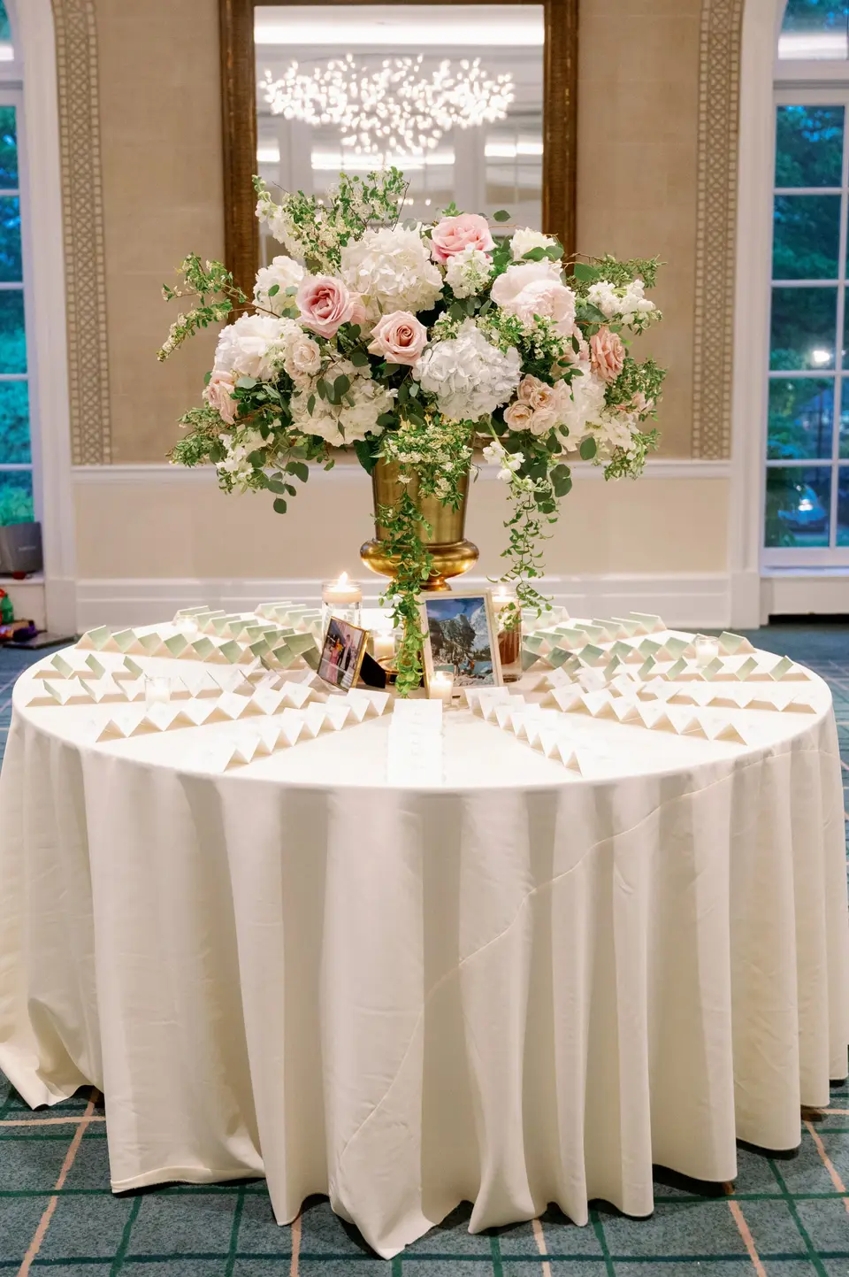 A round table of escort cards beneath a tall arrangement of pink and white flowers in a gold urn.