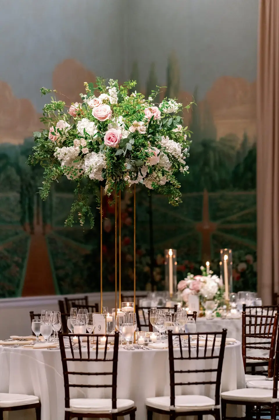 A tall arrangement of pink roses and greenery on a gold stand above a candlelit table, a painted garden mural behind.
