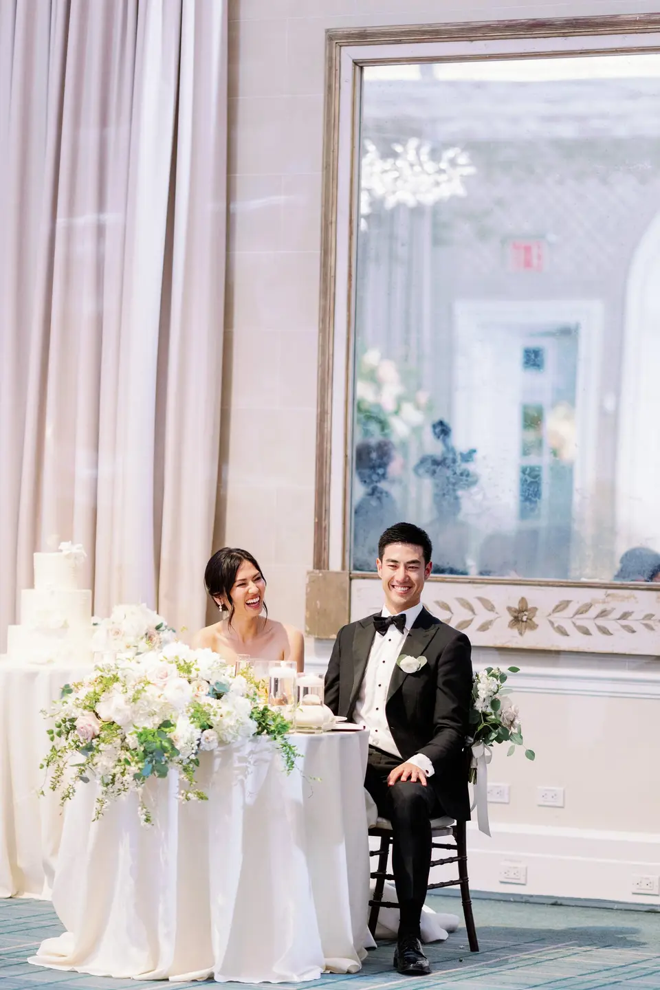 The bride and groom smile at the sweetheart table, white flowers spilling from its front.