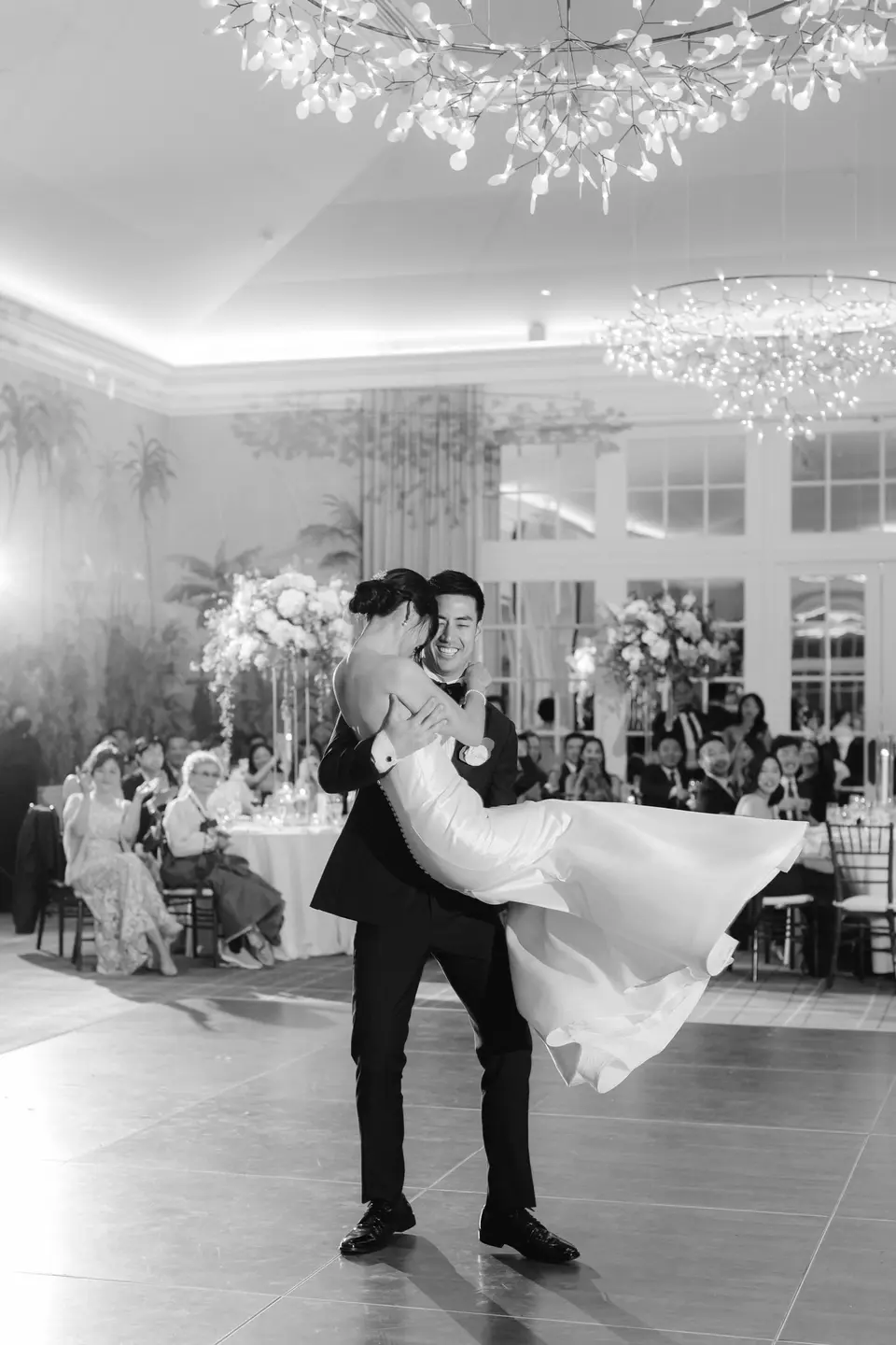 The groom lifts the bride off her feet on the dance floor beneath the chandeliers. Black and white.