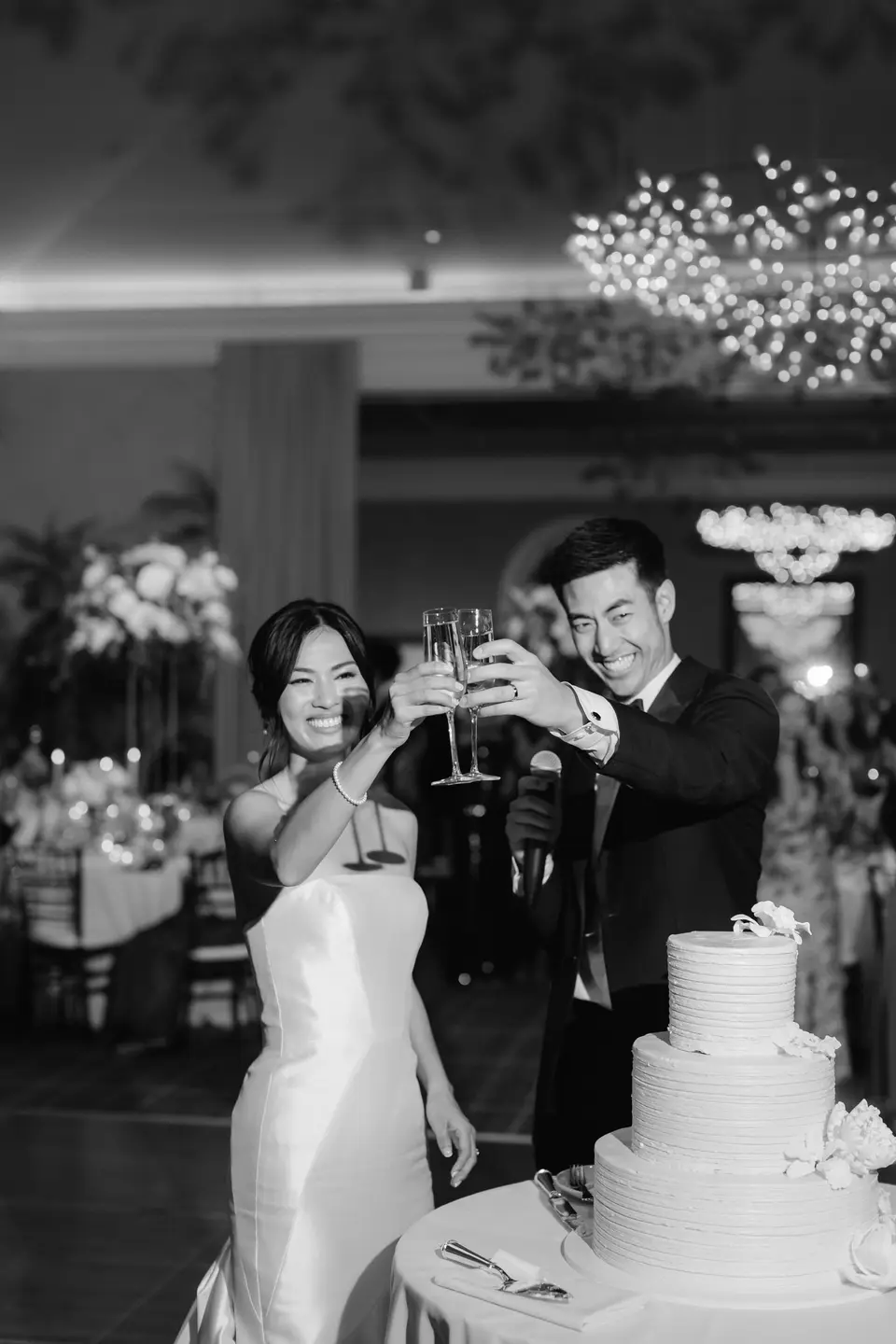 The bride and groom raise their champagne glasses beside the wedding cake. Black and white.