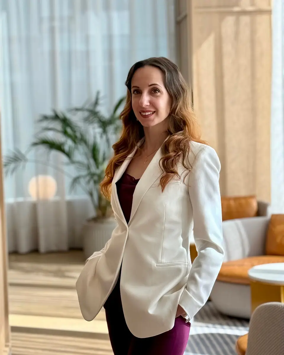 Olha Barabash smiling in a cream blazer and plum trousers, in a bright lounge with a palm and warm timber panelling.