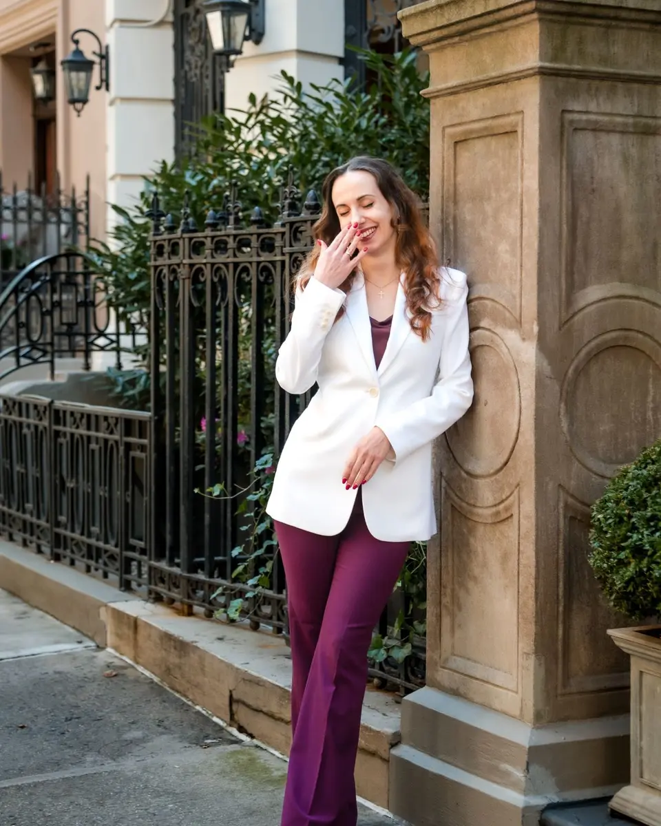 Olha Barabash laughing, a hand at her face, in a white blazer and plum trousers, leaning on a stone gatepost outside a New York townhouse.