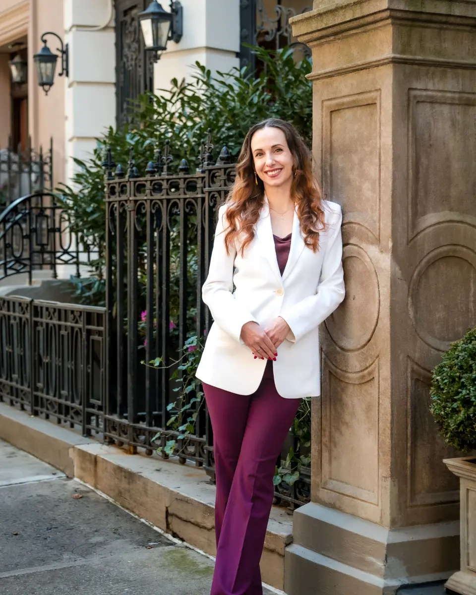 Olha Barabash smiles, leaning against a carved stone gatepost in a white blazer and plum trousers, a wrought iron fence and townhouse lanterns behind her.