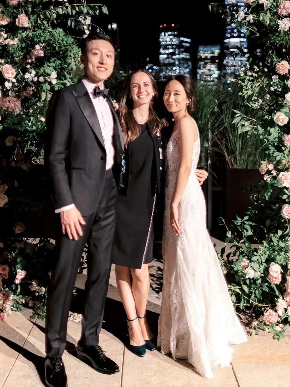 Olha smiling between a groom in a black tuxedo and a bride in a lace gown, in front of two tall arrangements of greenery and blush roses at night, with the Lower Manhattan skyline lit up behind them.