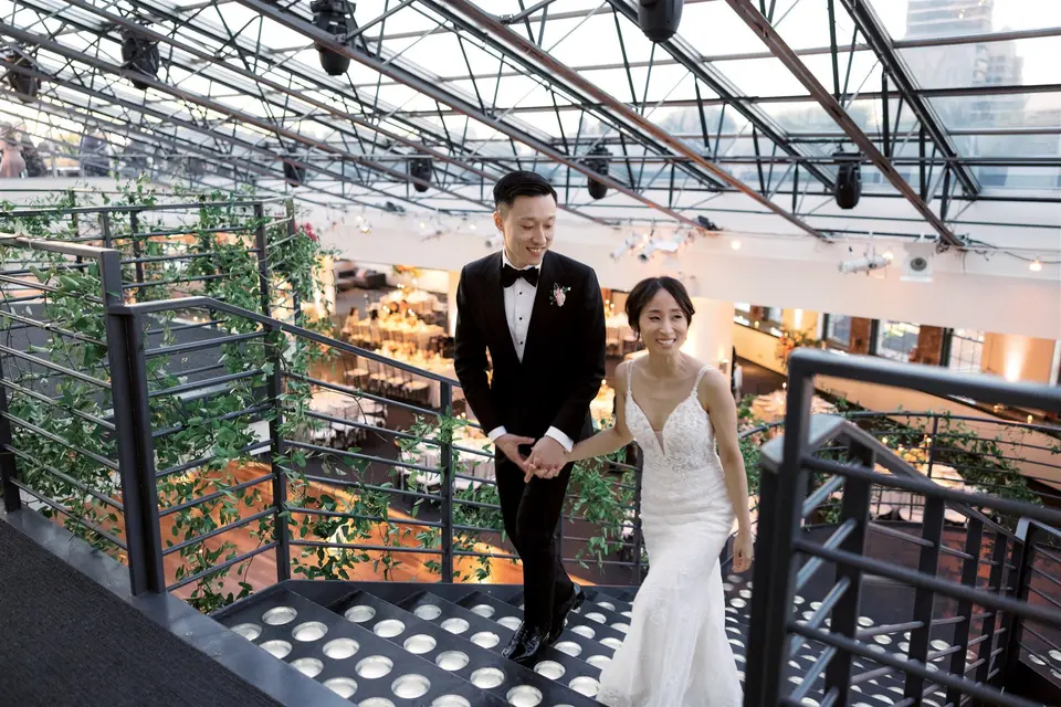 The couple climb a black iron staircase beneath a glass roof, the groom looking back at his bride.