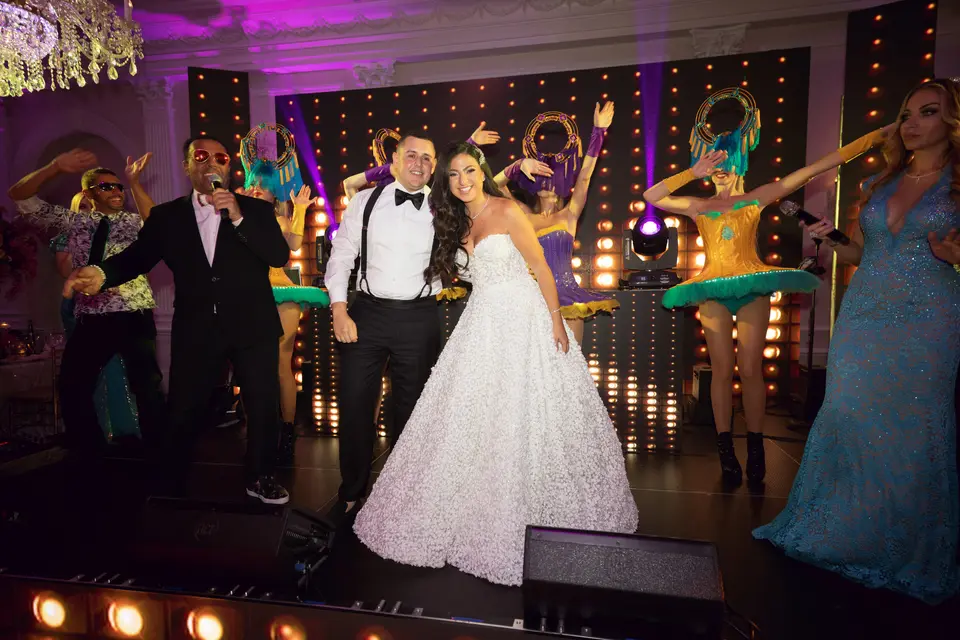 The couple dance on stage between performers in feathered costumes, the room lit in violet.